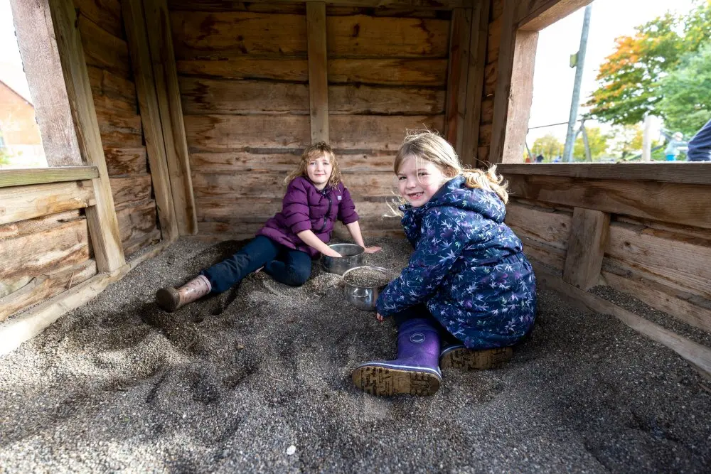 To piger leger i sandkassen under et halvtag af træ på skolens legeplads
